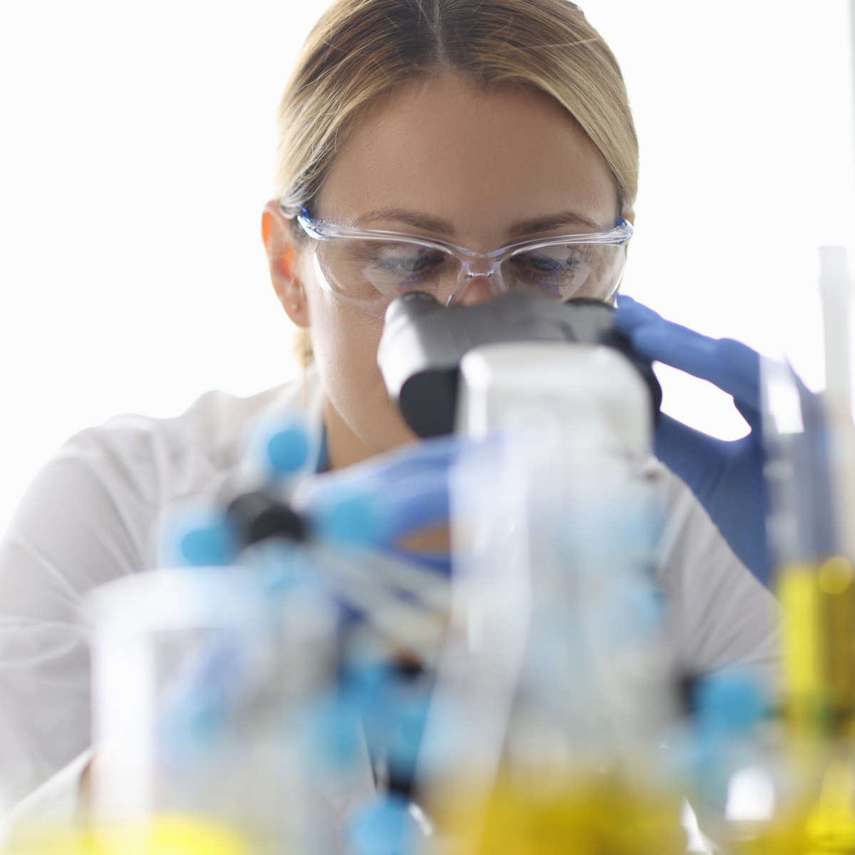 Researcher looks through microscope next to flask with yellow liquid. Oil quality control laboratory analysis concept